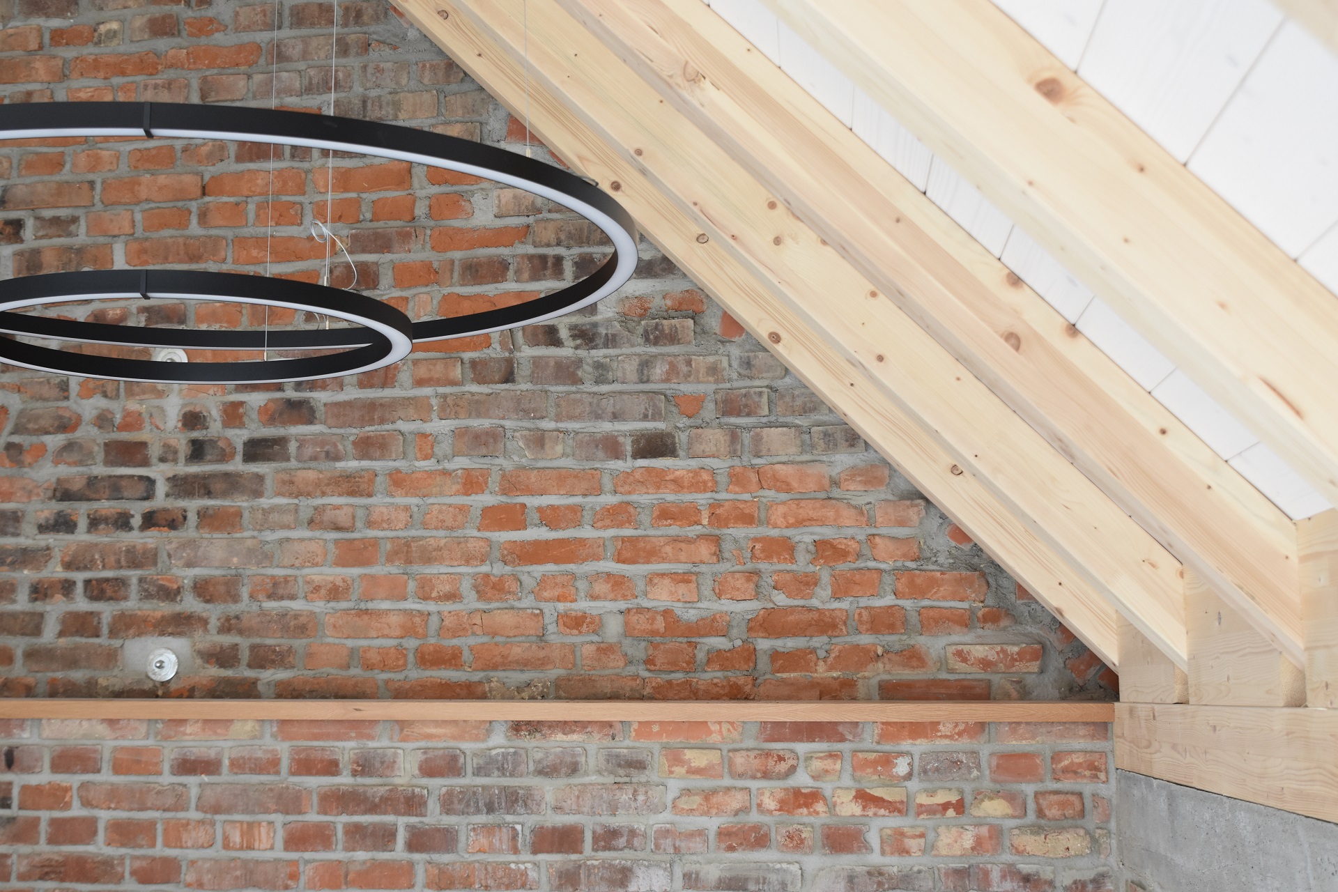 Dachstuhlausbau im modernen Loft-Stil mit einer Sichtziegelwand aus roten Backsteinen und sichtbaren Dachbalken aus hellem Nadelholz. Unter den weiß verkleideten Dachschrägen hängen zwei minimalistische, ringförmige LED-Pendelleuchten in Mattschwarz, die einen grafischen Kontrast zu den rustikalen Oberflächen bilden
