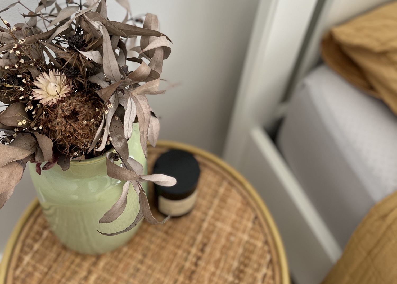 Detailaufnahme eines Nachttisches aus Rattan-Geflecht neben einem weiß gerahmten Bett mit senfgelber Bettwäsche. Auf dem Tisch sorgt ein Arrangement aus getrockneten Eukalyptusblättern und Strohblumen in einer hellgrünen Keramikvase sowie eine Duftkerze für ein harmonisches Schlafzimmer-Styling im Boho-Minimalismus