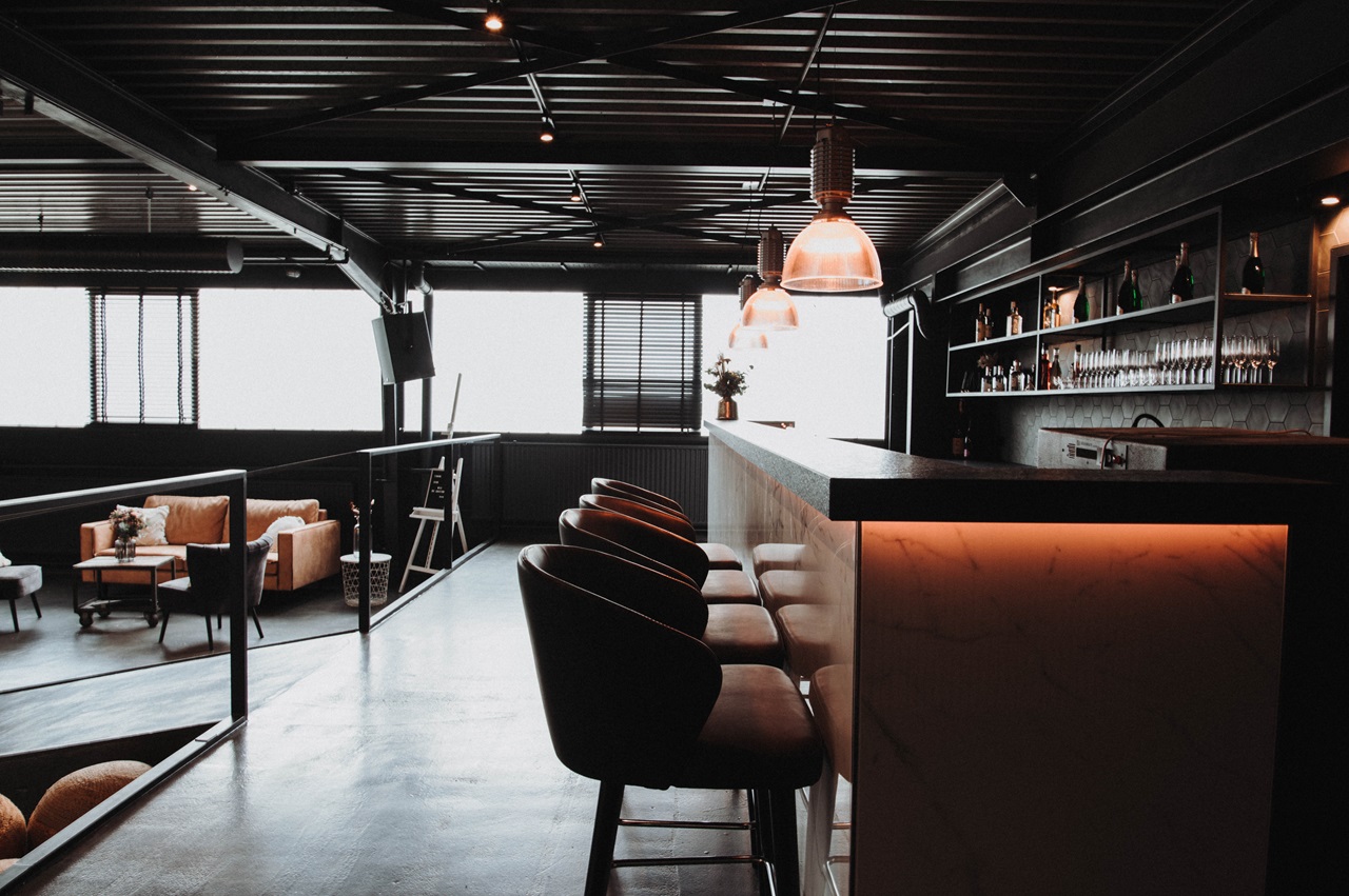 Industrial-Lounge auf zwei Ebenen mit einer beleuchteten Marmorbar und modernen Barhockern aus Leder im Vordergrund. Der obere Barbereich besticht durch eine offene, dunkle Deckenkonstruktion und kupferfarbene Hängeleuchten, während im tiefer gelegenen Hintergrund eine gemütliche Sitzecke mit Ledersofa erkennbar ist