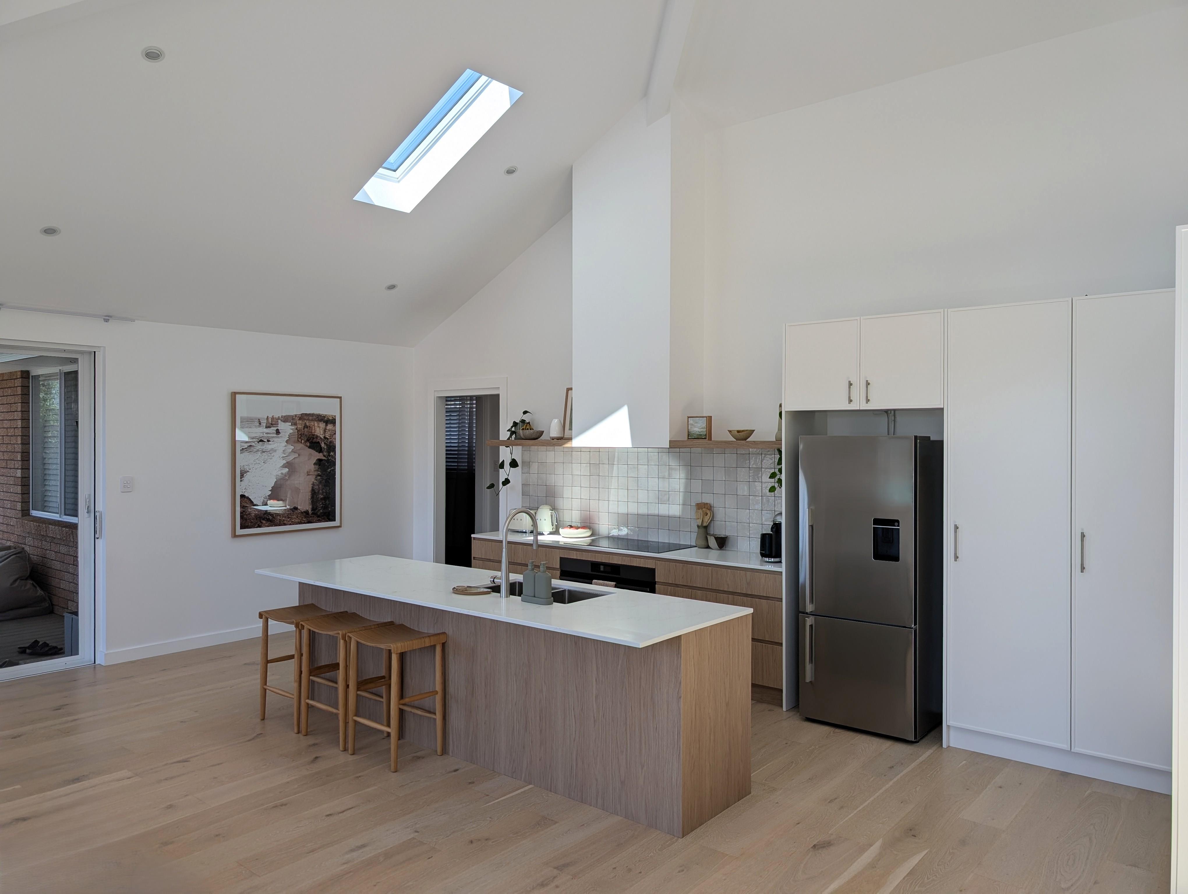 Lichtdurchflutete, offene Wohnküche im skandinavischen Design mit einer zentralen Kochinsel aus Eichenholz und integrierten Barhockern. Die Küchenzeile unter der hohen Deckenkante kombiniert minimalistische weiße Hochschränke, einen Edelstahl-Kühlschrank, handgefertigte quadratische Wandfliesen und ein großes Dachfenster, das natürliches Tageslicht auf den hellen Holzboden wirft