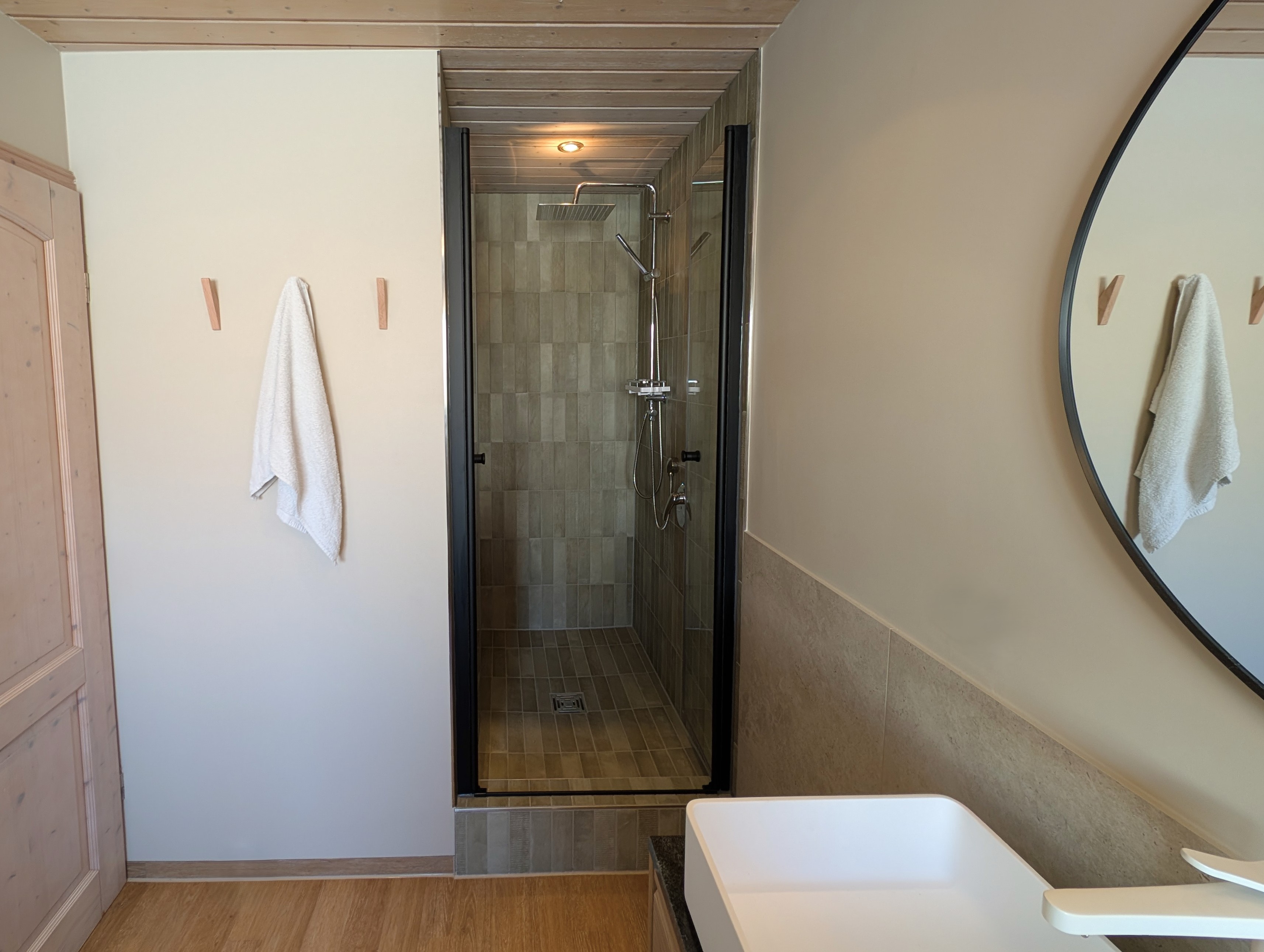 Minimalistisches Badezimmer mit einer Duschnische, die mit vertikal verlegten, salbeigrünen Riemchenfliesen und einer schwarzen Glastür gestaltet ist. Im Vordergrund ergänzen ein weißes Aufsatzwaschbecken, ein großer runder Spiegel und eine Wandfläche mit schlichten Holz-Handtuchhaken vor einer hellen Holzdecke das moderne Spa-Konzept