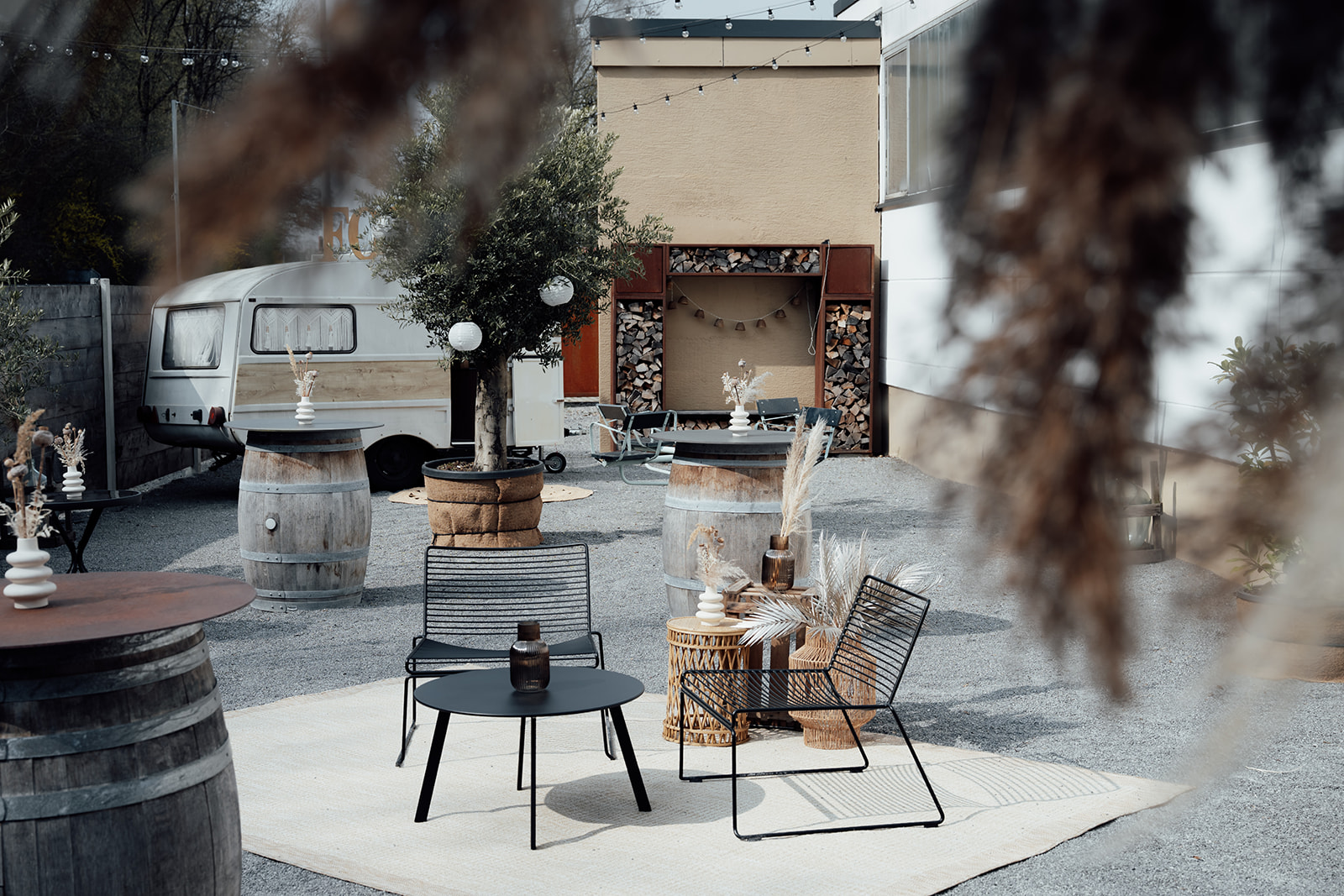 Moderner Outdoor-Loungebereich im Boho-Industrial-Stil auf einem grauen Kiesplatz mit minimalistischen Drahtgitter-Sesseln auf einem hellen Teppich. Das Gestaltungskonzept nutzt alte Weinfässer als Stehtische, einen Olivenbaum im Jutesack, ein Holzlager als architektonischen Raumteiler und einen restaurierten Vintage-Wohnwagen im Hintergrund