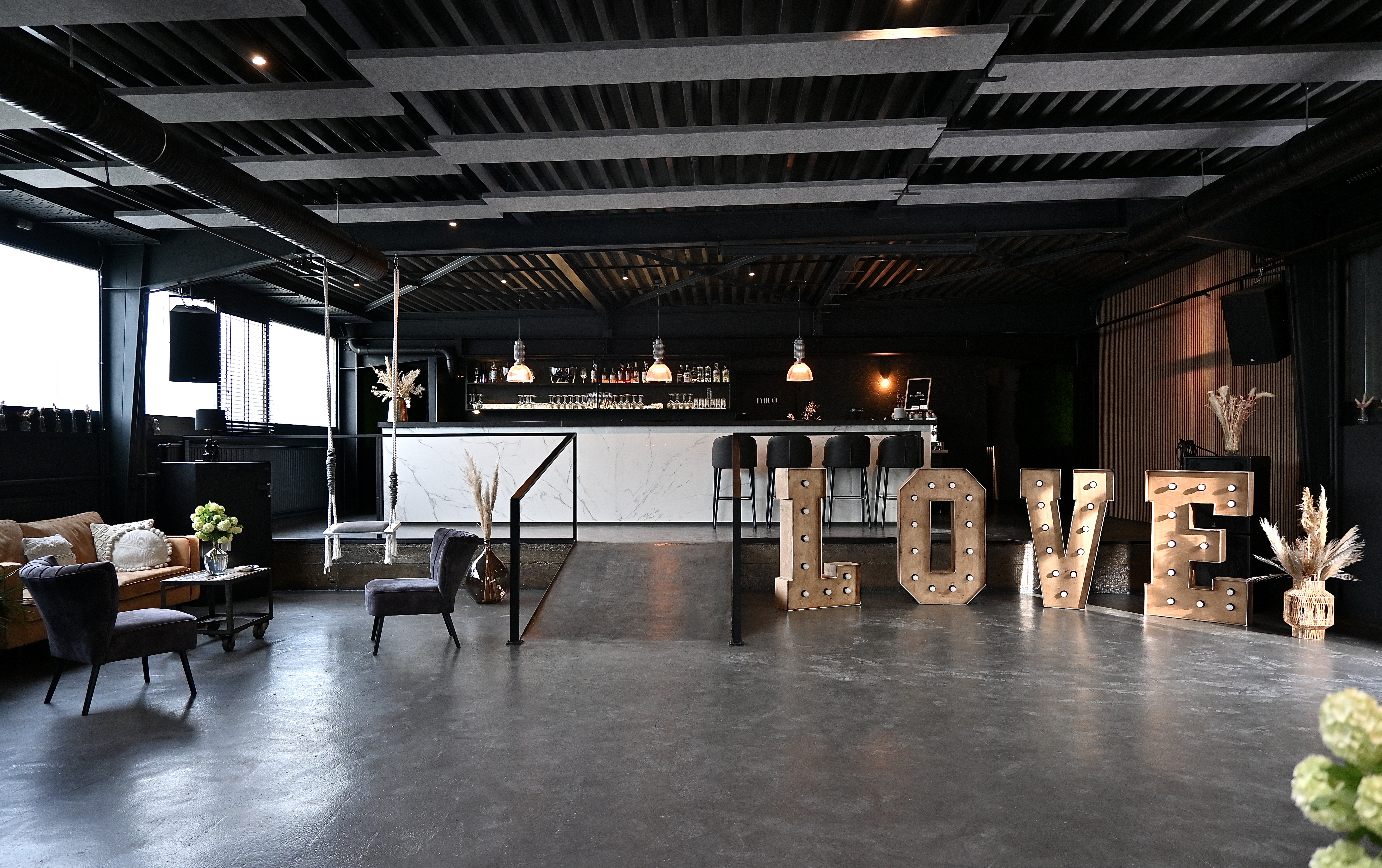 Großzügiges Event-Interieur im Industrial-Style mit geschliffenem Betonboden, einer Marmorbar im Hintergrund und großen, beleuchteten „LOVE“-Leuchtbuchstaben aus Holz. An der dunklen Deckenstruktur sind graue Akustiksegel montiert, während links eine Sitzecke mit Ledersofa und rechts eine vertikale Holzlattenwand das Raumkonzept abrunden