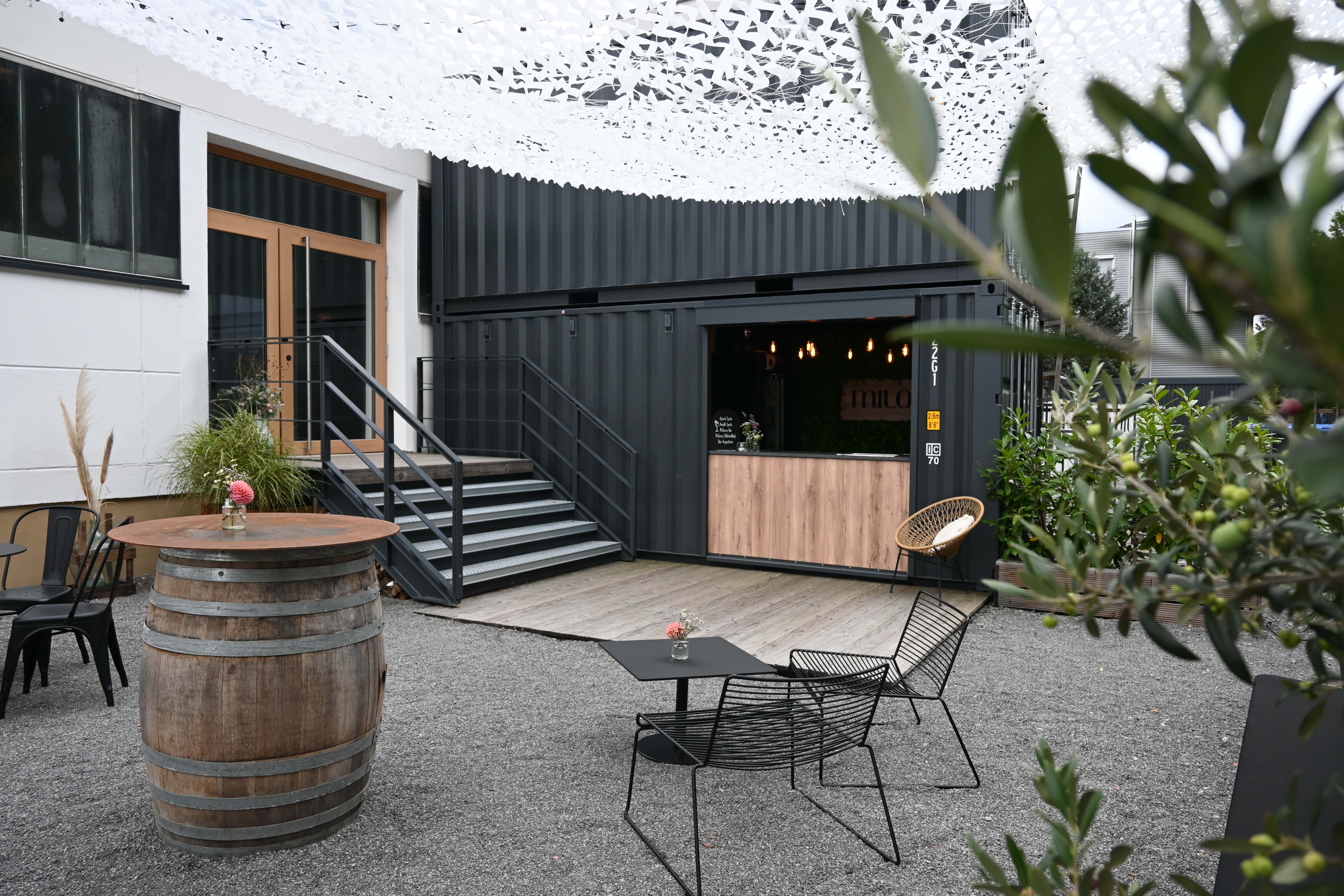 Moderner Außenbereich im Industrial-Design mit einer Kiosk-Bar aus umgebauten, anthrazitfarbenen Schiffscontainern und einer Holzterrasse. Der gestaltete Kiesplatz im Vordergrund verfügt über minimalistische Drahtsessel, Weinfässer als Stehtische und ein weißes Sonnensegel in Camouflage-Optik für eine strukturierte Beschattung