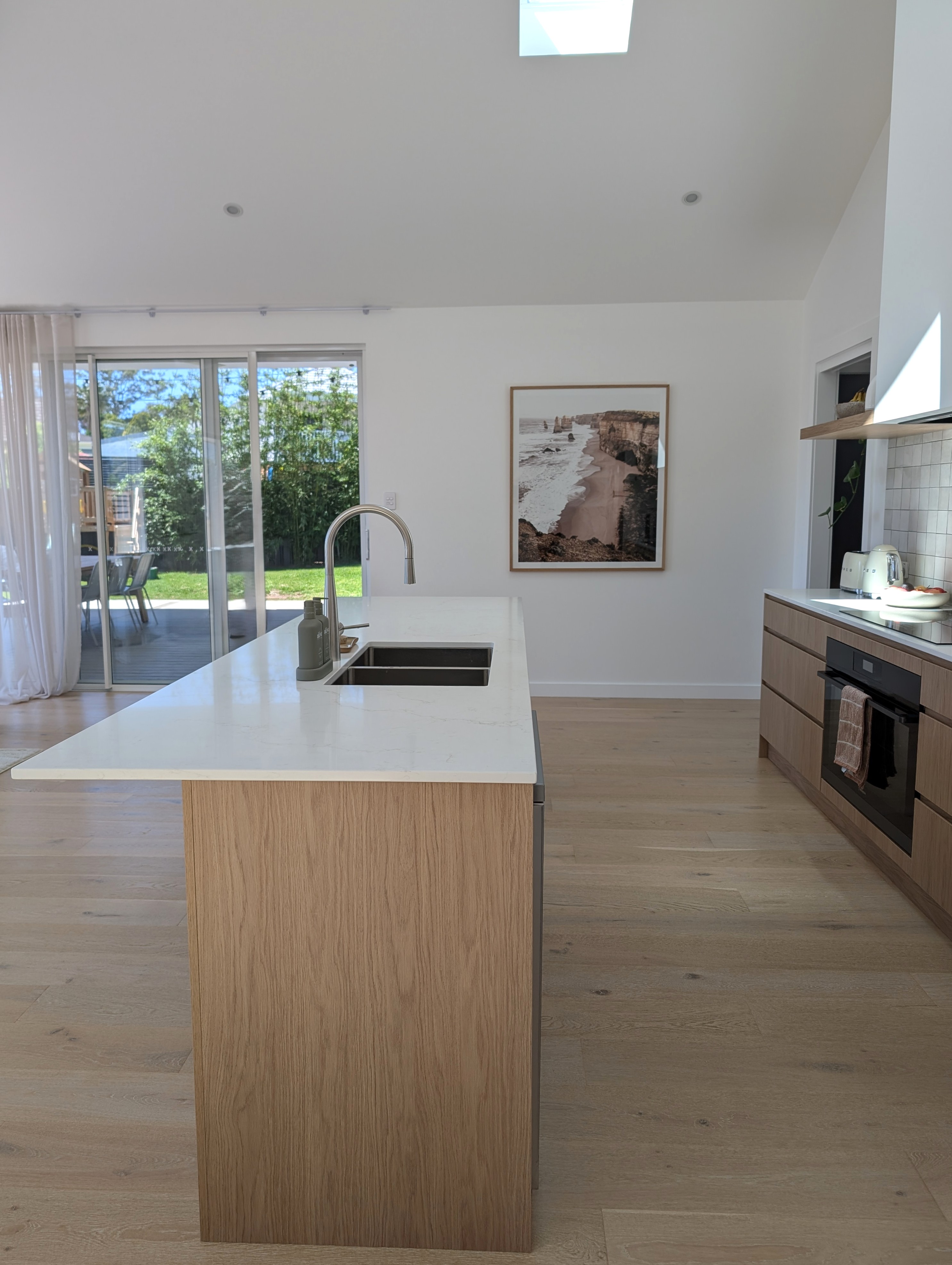 Moderne Wohnküche mit Blick auf eine Kücheninsel aus hellem Eichenholz mit weißer Quarz-Arbeitsplatte, Spüle und bogenförmiger Edelstahl-Armatur. Im Hintergrund öffnet eine große Glasschiebetür den Raum zum Garten, während ein gerahmtes Landschaftsbild an der weißen Wand und die grifflose Küchenzeile rechts das minimalistische Raumdesign unterstreichen