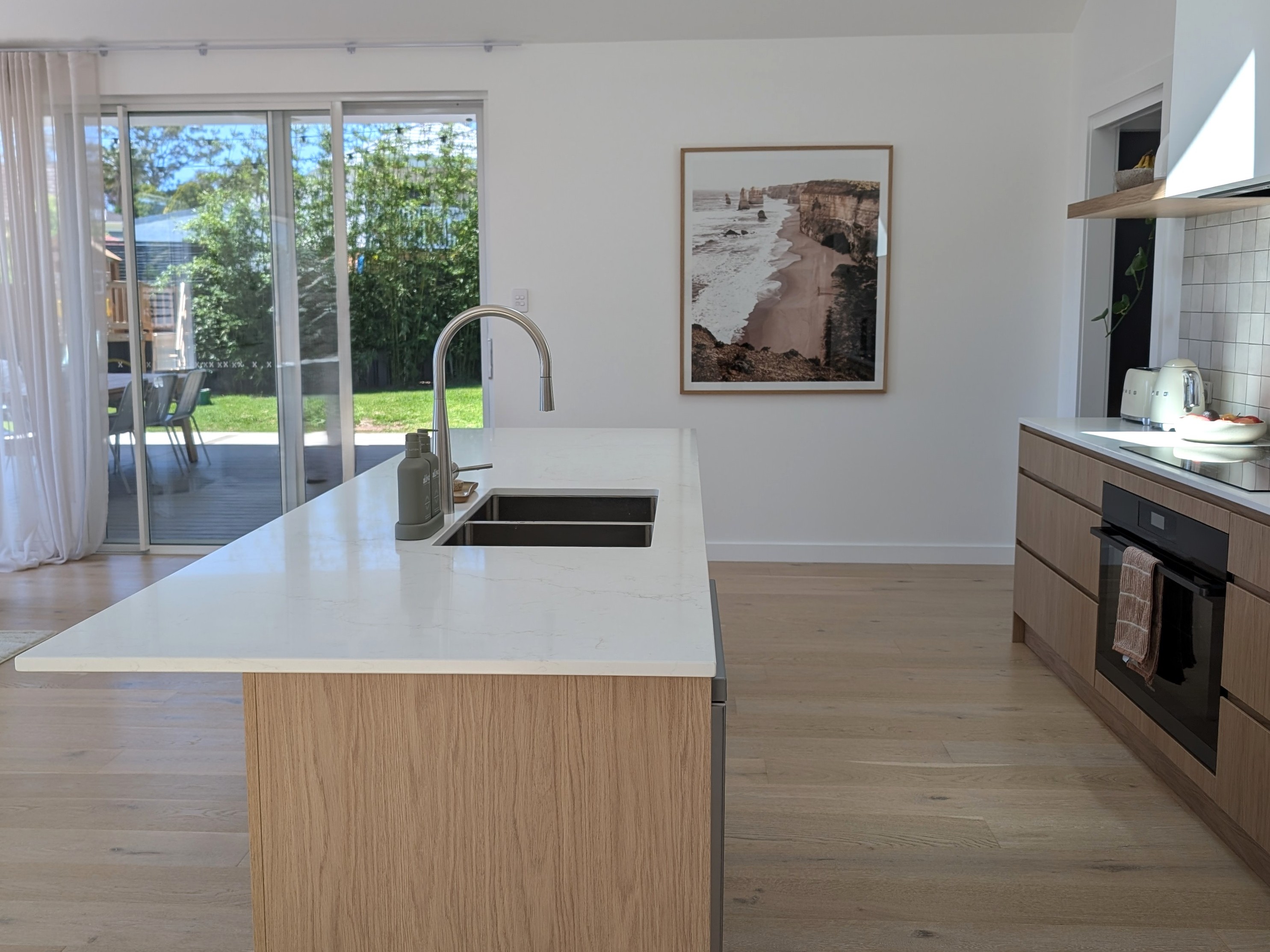 Moderne Wohnküche mit Blick auf eine Kücheninsel aus hellem Eichenholz mit weißer Quarz-Arbeitsplatte, Spüle und bogenförmiger Edelstahl-Armatur. Im Hintergrund öffnet eine große Glasschiebetür den Raum zum Garten, während ein gerahmtes Landschaftsbild an der weißen Wand und die grifflose Küchenzeile rechts das minimalistische Raumdesign unterstreichen