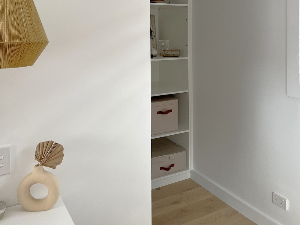Blick durch eine Wandöffnung auf ein integriertes, weißes Einbauregal in einer Nische mit hellem Holzboden. Im linken Vordergrund setzen eine gewebte Hängeleuchte aus Bast, eine beige Keramikvase mit Palmenblatt-Deko und minimalistische Aufbewahrungsboxen mit Lederschlaufen warme, skandinavische Akzente vor cleanen weißen Wänden
