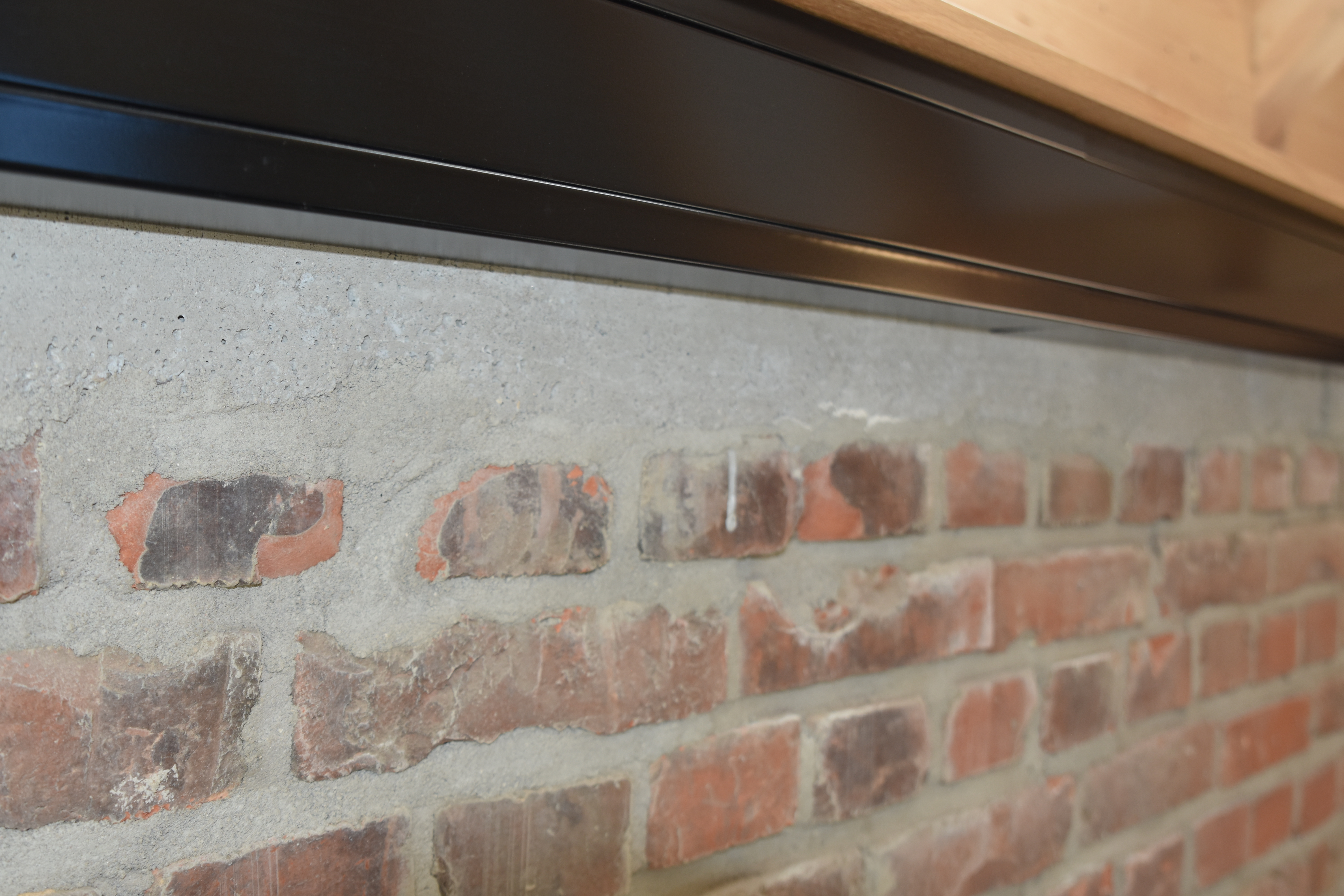 Architektonische Detailaufnahme eines Wandanschlusses mit einer rustikalen Backsteinwand und einer darüber liegenden Sichtbetonkante. Das Konstruktionsdetail im Industrial-Stil wird durch ein minimalistisches, mattschwarzes Metallprofil und eine darüber ansetzende Verkleidung aus hellem Naturholz sauber abgeschlossen