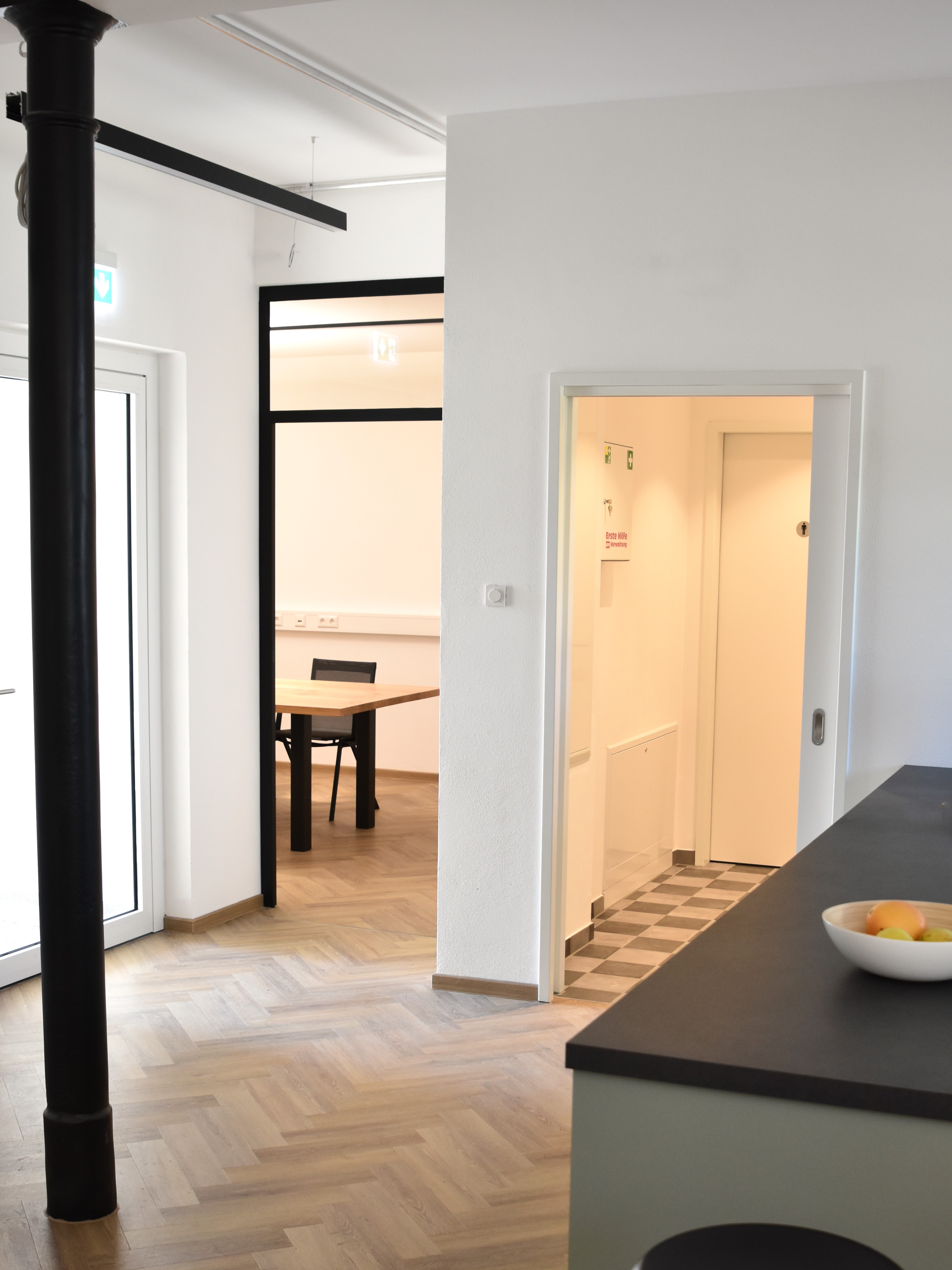 Moderner Büro- und Empfangsbereich mit Blick auf einen Konferenzraum und sanitäre Anlagen. Das Design kombiniert einen edlen Parkettboden im Fischgrätmuster mit einer dunklen Küchentheke im Vordergrund, einer markanten schwarzen gusseisernen Rundsäule und einer verglasten Trennwand im Loft-Stil mit schwarzem Stahlrahmen