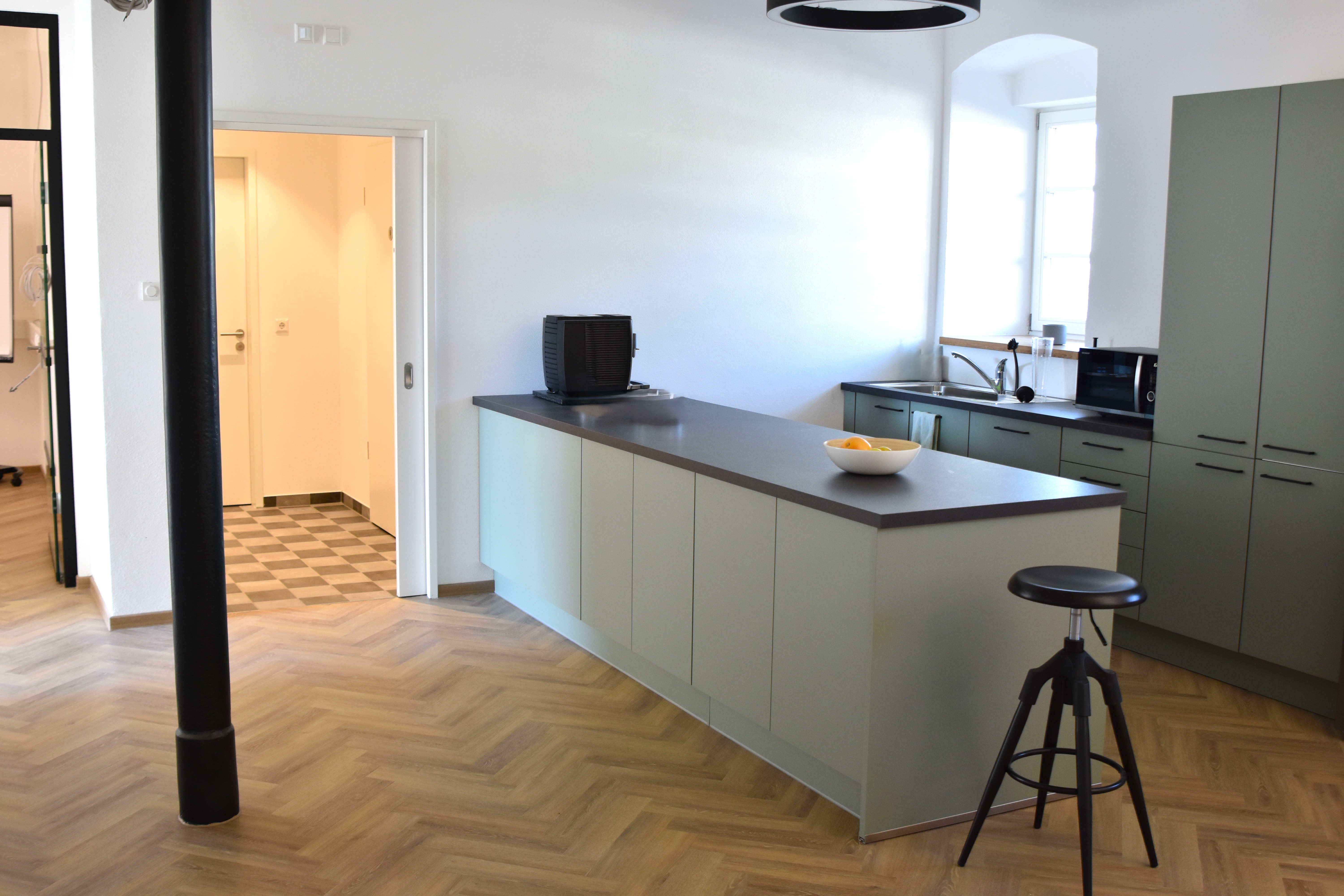 Moderne Büroküche mit einer freistehenden Kücheninsel in Salbeigrün und dunkelgrauer Arbeitsplatte sowie einem passenden Barhocker. Das Raumkonzept verbindet grifflose, olivgraue Hochschränke mit einer integrierten Küchenzeile am Fenster, einem edlen Fischgrätparkett und einer markanten schwarzen Gusseisensäule im Vordergrund
