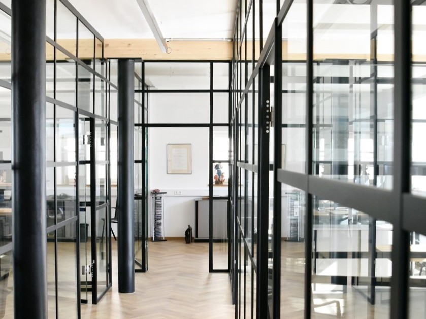 Modernes Bürokonzept mit transparenten Trennwänden im Loft-Stil aus schwarzen Lofttüren-Stahlrahmen und Glaselementen. Das Raumdesign kombiniert einen edlen Parkettboden im Fischgrätmuster mit industriellen, schwarzen Rundsäulen und sichtbaren Holzbalken an der Decke, die für eine helle und strukturierte Arbeitsatmosphäre sorgen