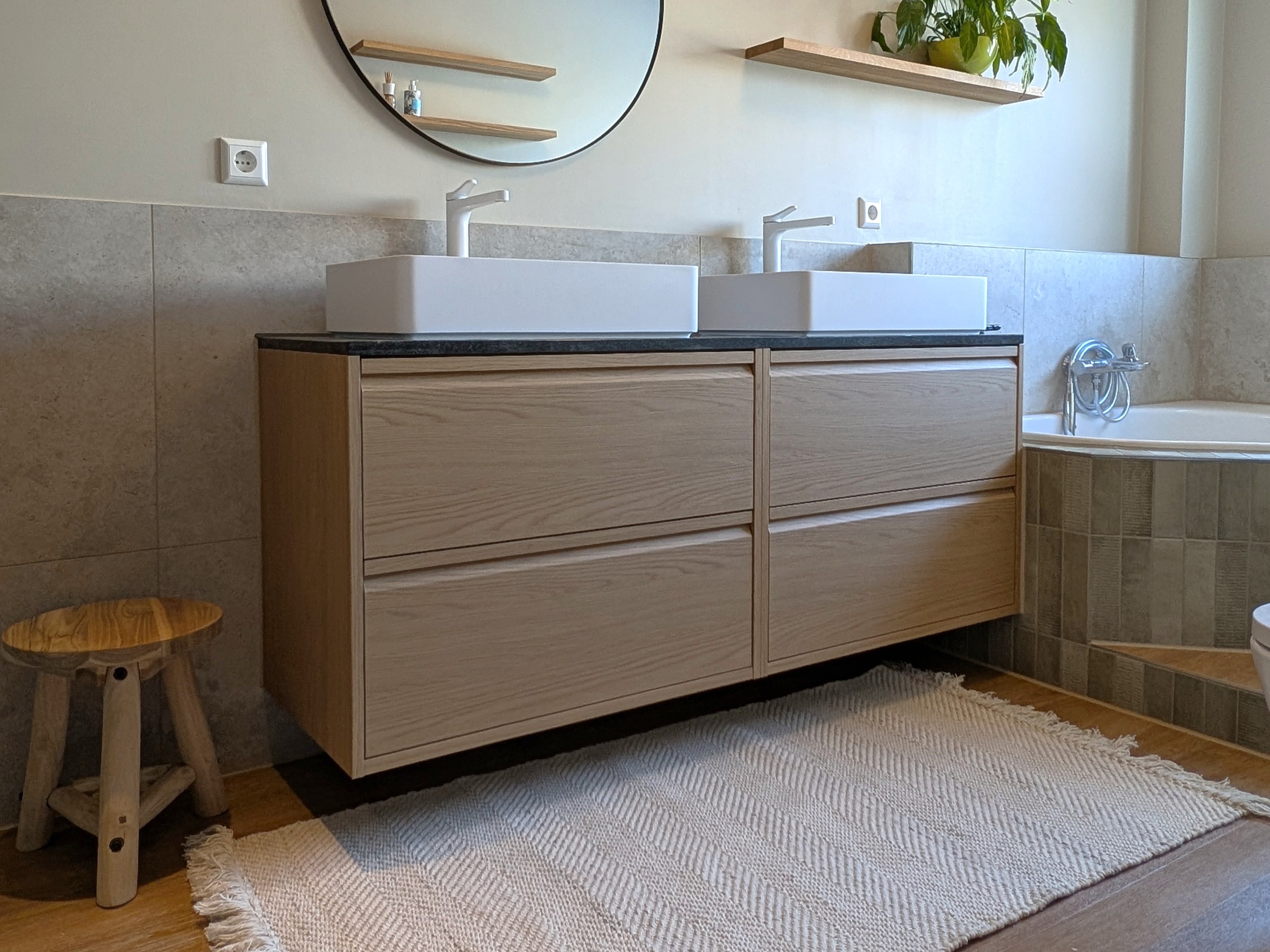 Detailansicht eines schwebenden Doppelwaschtischs aus hellem Eichenholz mit integrierten Griffleisten und einer dunklen Naturstein-Arbeitsplatte. Auf dem Möbel sitzen zwei minimalistische, rechteckige Aufsatzwaschbecken, ergänzt durch einen runden Spiegel, einen gewebten Teppichläufer mit Fransen und einen rustikalen Holzhocker auf dem warmen Holzboden